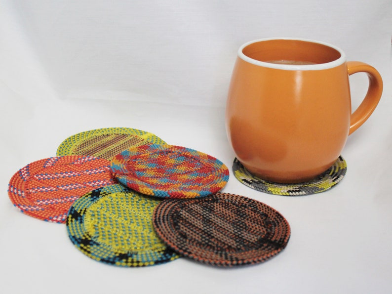 May include: A round orange mug with a white rim sits on a circular coaster with a grey and yellow pattern. Four additional round coasters are arranged nearby, featuring woven designs in vibrant colors including orange, yellow, blue, and black.