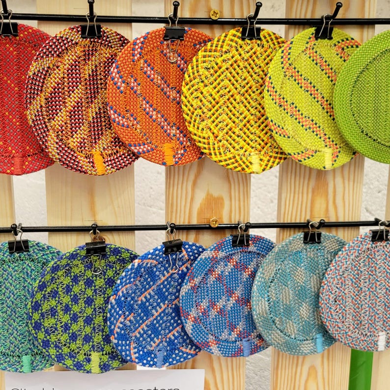 May include: A collection of twelve circular woven coasters displayed in two rows, featuring a variety of vibrant colors including red, orange, yellow, lime green, blue, and teal, each secured with a black metal binder clip on a wooden rack.