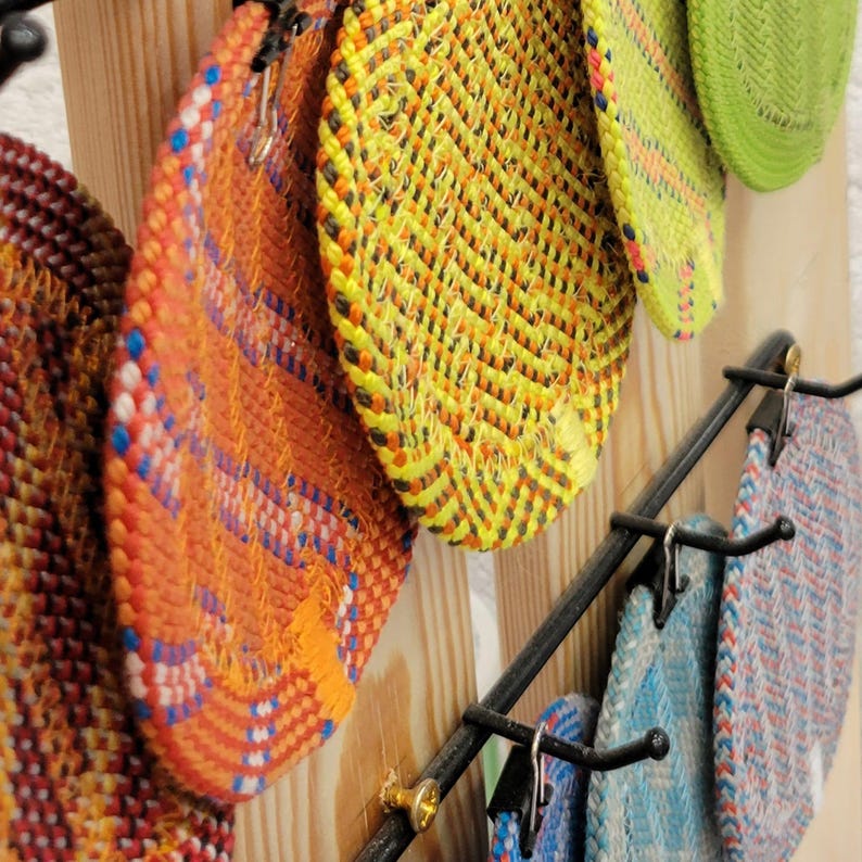 May include: A collection of circular woven coasters in various colors including orange, yellow, lime green, and blue, featuring a herringbone pattern. The coasters are suspended from metal hooks attached to a vertical light-colored wooden board.