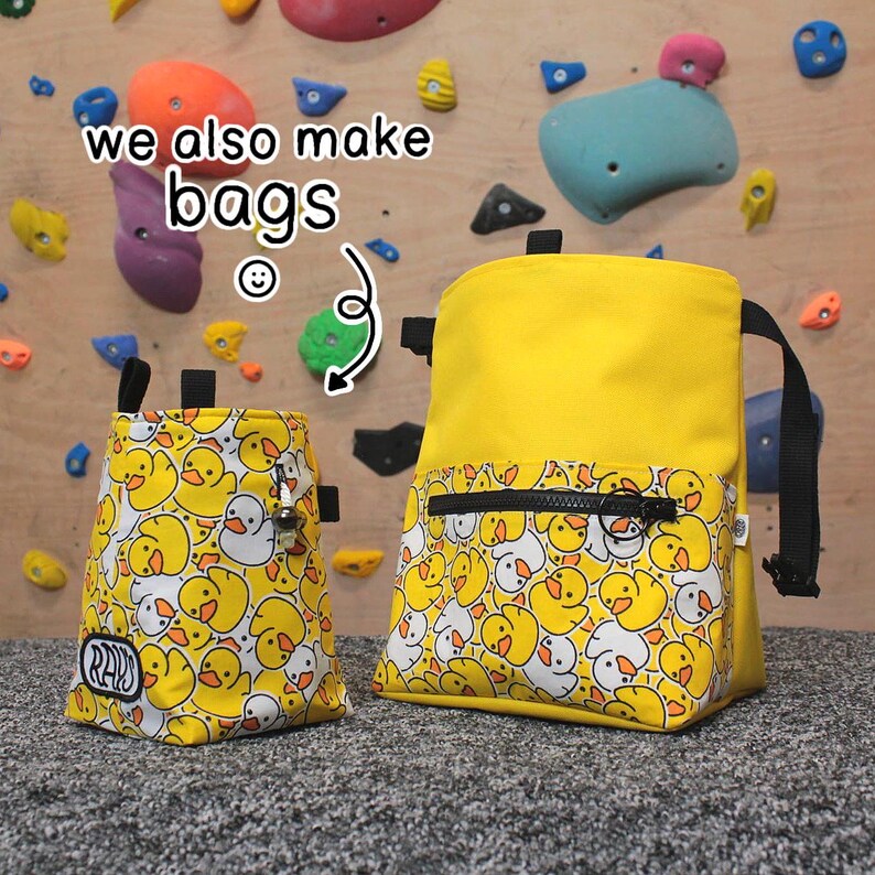Pode incluir: Duas bolsas de giz de escalada amarelas com um padr&atilde;o de pato branco. A bolsa maior tem um bolso com z&iacute;per e uma al&ccedil;a preta. A bolsa menor tem uma al&ccedil;a preta e um mosquet&atilde;o de metal. O texto "we also make bags" &eacute; vis&iacute;vel na imagem.
