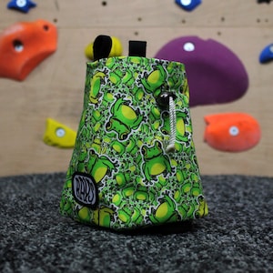 May include: A green and white climbing chalk bag featuring a frog design. It has a drawstring closure and a black loop. The bag is patterned with cartoon frogs and has the text "Rays" on the front.