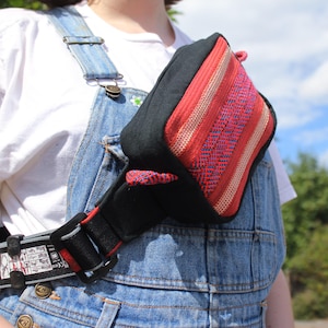 Puede incluir: Un bolso de cintura negro y rojo con un diseño tejido a rayas. La bolsa se lleva cruzada sobre el pecho, asegurada por una correa negra con un detalle rojo. El bolso de cintura se combina con un peto vaquero y una camiseta blanca.