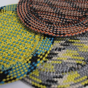 May include: Three circular coasters featuring a woven texture. One coaster displays a bright yellow and blue grid pattern, another features a black and orange spiral weave, and the third shows a grey, yellow, and white geometric pattern.