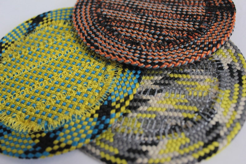 May include: Three circular coasters featuring a woven texture. One coaster displays a bright yellow and blue grid pattern, another features a black and orange spiral weave, and the third shows a grey, yellow, and white geometric pattern.