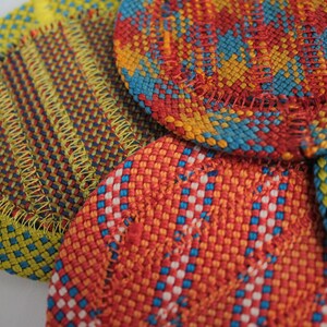 May include: A collection of circular woven coasters featuring intricate patterns in vibrant colors including yellow, red, blue, and orange. Each coaster is finished with a visible stitched border, showcasing a dense, textured weave of interlaced threads.