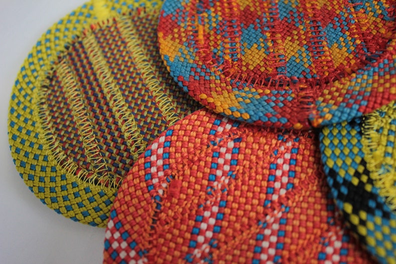 May include: A collection of circular woven coasters featuring intricate patterns in vibrant colors including yellow, red, blue, and orange. Each coaster is finished with a visible stitched border, showcasing a dense, textured weave of interlaced threads.