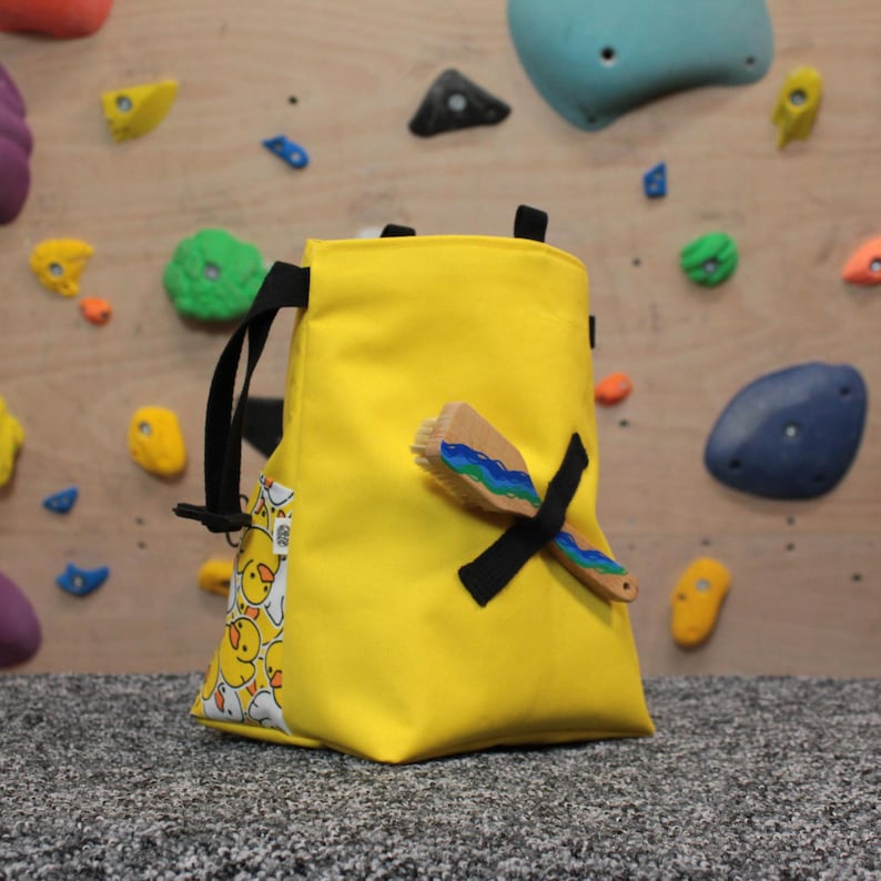 Pode incluir: Uma bolsa de giz de escalada amarela com uma al&ccedil;a preta e uma escova com listras azuis e verdes. A bolsa tem um pequeno bolso com um padr&atilde;o de pintinhos amarelos.