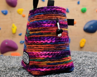 May include: A colorful, knitted climbing chalk bag with a drawstring closure and a small black loop. The bag features a vibrant mix of purple, orange, pink, and green horizontal stripes. A small patch with the word "RAYS" is visible.