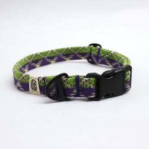 May include: A woven pet collar featuring a green and purple plaid pattern with a black plastic side-release buckle, a black metal D-ring, and a small off-white fabric tag with the text RAW printed in a circular design on the strap.