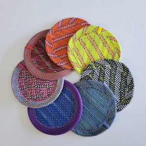 May include: A collection of seven circular coasters arranged in a fan shape. Each coaster features a distinct woven pattern with vibrant colors including orange, neon yellow, black and white, blue, purple, and multi-colored pink and blue threads.