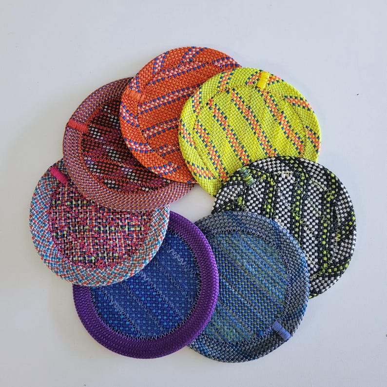 May include: A collection of seven circular coasters arranged in a fan shape. Each coaster features a distinct woven pattern with vibrant colors including orange, neon yellow, black and white, blue, purple, and multi-colored pink and blue threads.