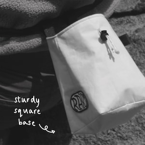 May include: White chalk bag with a black logo that says "RAD" and a sturdy square base. The bag is hanging from a person's belt.