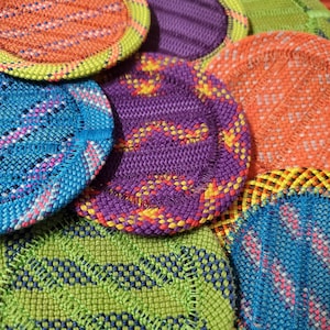 May include: A collection of circular, woven fabric coasters featuring vibrant, multi-colored geometric patterns in shades of orange, purple, blue, and lime green. Each coaster has a distinct textured weave and a stitched border edge.