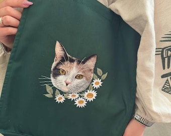 Può includere: Una borsa tote verde scuro con un ricamo di un volto di gatto circondato da margherite bianche e foglie verdi. Il gatto ha un pelo marrone, nero e bianco con occhi gialli. La borsa ha un design semplice con due manici.