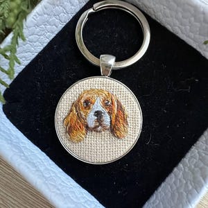 Puede incluir: Un llavero circular que presenta un bordado detallado de la cara de un perro marrón y blanco sobre un fondo de tela beige. El bordado está colocado dentro de un marco de metal plateado y unido a una anilla de metal plateado estándar.