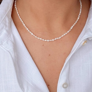 Könnte beinhalten: Eine zarte Perlenkette aus kleinen, weißen Perlen. Die Kette wird vor einem weißen Button-Down-Hemd getragen. Die Perlen sind oval geformt und gleichmäßig angeordnet, was einen klassischen und eleganten Look erzeugt.