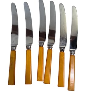 May include: Set of six vintage butter knives. Each knife features a stainless steel blade and a handle in a warm, amber-colored hue. The handles have a rectangular shape. The knives are arranged on a white background.
