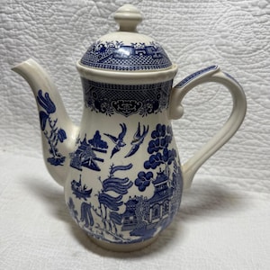 May include: Cream-colored ceramic teapot with a blue Willow pattern. The teapot features a lid, spout, and handle. The blue design includes pagodas, birds, and floral elements. A classic piece for serving tea.