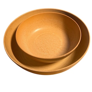 May include: Two nested, light brown bowls. The larger bowl has a wide, shallow shape, while the smaller bowl is rounder and fits inside. Both bowls have a textured surface and a warm, earthy tone, suggesting a natural material.