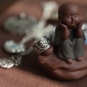 May include: A small, brown ceramic figurine of a meditating person with closed eyes, sitting on a brown base. The figurine is surrounded by silver jewellery, including a ring with a detailed design.