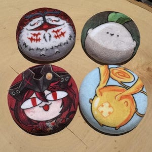 Può includere: Quattro spille rotonde in peluche con disegni di personaggi dei cartoni animati. Le spille presentano una varietà di colori, tra cui rosso, bianco, grigio e giallo. Le spille sono disposte su una superficie di legno.