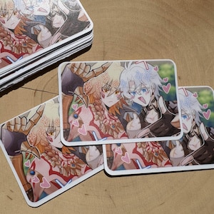 以下が含まれることがあります： 2人のキャラクターのアニメ風イラストが描かれた長方形のステッカーの束と、いくつかのバラバラのステッカー。キャラクターは明るい髪をしており、ピンクのハートに囲まれています。ステッカーは木製の表面にあります。