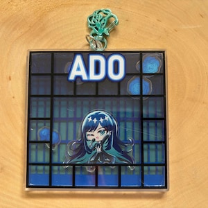 May include: A square acrylic charm featuring a cartoon illustration of a character with long blue hair and teal accents. The charm has the word "ADO" in white block letters. A teal-colored charm loop is attached at the top.