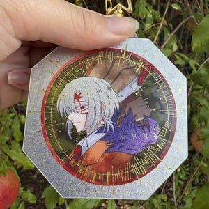 May include: An octagonal acrylic charm featuring a silver glitter border and a gold star-shaped clasp. The center displays an anime-style illustration of two characters with white and purple hair, surrounded by a circular red frame with gold line art.