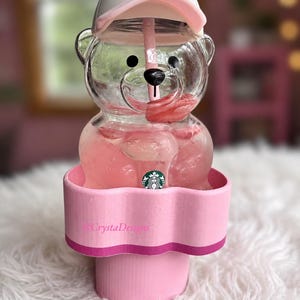 以下が含まれることがあります： ピンク色のスターバックスのキャップ型リッドとストローが付いた、透明なクマの形のタンブラー。クマには黒い鼻と目があり、胸には小さなスターバックスのロゴが付いています。波状の縁を持つピンク色のカップホルダーに入っています。