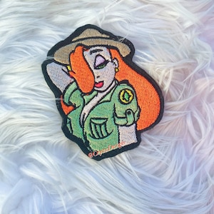 Op de afbeelding: Geborduurde patch met een cartoonvrouw met rood haar, gekleed in een groen uniform en een beige hoed. De patch heeft een zwarte rand en bevat de tekst "@CrystalDesigns".