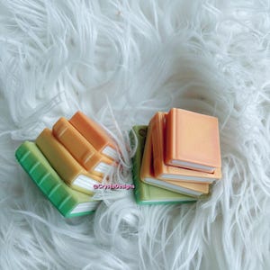 May include: Miniature books in shades of green, yellow, and orange, with white pages, are arranged on a fluffy white surface. The books are stacked and scattered, creating a playful and colorful display. The text "@CrystaDesigns" is visible.