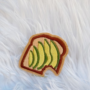 May include: A brown and tan embroidered patch of a slice of bread with avocado slices on top. The text "@CrystaDesigns" is embroidered on the bottom of the patch.