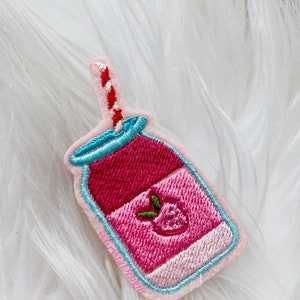 Puede incluir: Parche bordado para planchar de un batido de fresa rosa y rojo en un tarro de vidrio con una pajita rayada roja y blanca. El parche tiene un contorno azul claro y el texto "@kristadesigns" es visible en la esquina inferior derecha.