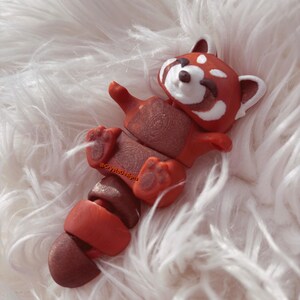 Op de afbeelding: Handgemaakte rode panda figuur met witte accenten, liggend op zijn rug. De figuur heeft een gedetailleerd ontwerp, inclusief een gesegmenteerde staart en poten. De achtergrond is een zachte, witte, pluizige textuur.