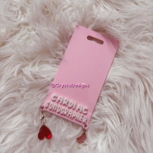 May include: A pink badge holder with the text "CARDIAC SONOGRAPHER" in white, featuring a heart charm and a charm with a face. The badge holder is on a fluffy white surface.