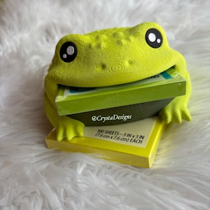 May include: A lime green 3D-printed frog figurine holding a stack of yellow sticky notes in its mouth. The notes are labeled 100 sheets-3 in x 3 in (7.6 cm x 7.6 cm) each. The frog has large black and white eyes and the text @CrystaDesigns.