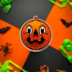 May include: A close-up of a Halloween-themed pumpkin sticker with a smiling face and wide eyes. The sticker is on a black and orange background with other Halloween decorations.