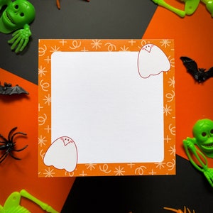 May include: A white card with an orange border and a repeating pattern of stars, plus signs, and swirls. Two cartoon ghosts are on the card, one in each corner. The card is surrounded by Halloween decorations, including spiders, skeletons, and bats.