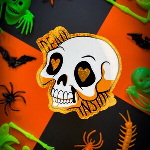 May include: A glittery sticker with a skull design and the words "Dead Inside" in black text. The skull has two hearts for eyes and a dripping tear. The sticker is on a black and orange background with various Halloween decorations.