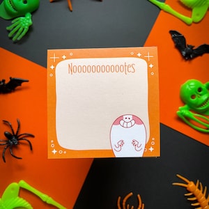 May include: Orange and white notepad with the text "Nooooooooootes" and a cartoon ghost with a smile. The notepad is surrounded by Halloween decorations, including spiders, skeletons, and bats.