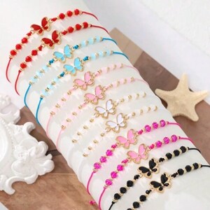May include: A collection of colorful butterfly charm bracelets. The bracelets feature red, blue, pink, and black cords, adorned with beads and butterfly pendants in various colors. The bracelets are displayed on a white surface.
