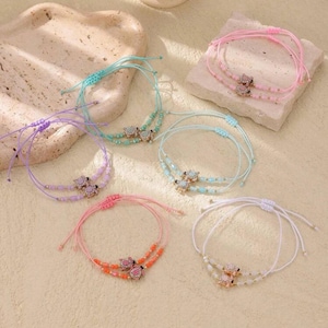 May include: A collection of colorful friendship bracelets. Each bracelet features a small, decorative charm and is adorned with beads. The bracelets are in shades of pink, blue, purple, and white, with adjustable string closures.