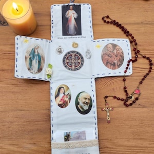 Pode incluir: Um objeto artesanal em forma de cruz com várias imagens religiosas e texto. A cruz apresenta imagens de Jesus, Maria e outros santos, juntamente com um rosário e uma vela acesa. O texto "Jésus, j'ai confiance en Vous" é visível.