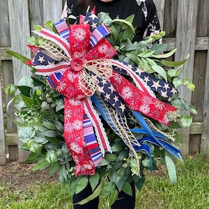 May include: A patriotic wreath featuring a large red, white, and blue bow with star and firework patterns. The bow is centered on a bed of green foliage. The wreath includes burlap and lace accents, and blue ribbon streamers.