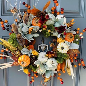May include: A fall-themed wreath with pumpkins, berries, and foliage in shades of orange, white, and green. The wreath includes a variety of textures, such as pinecones and dried grasses. A silver door knocker with the text "J & T Pagliaro" is visible.