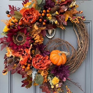 May include: A decorative autumn wreath featuring a grapevine base adorned with deep burgundy and vibrant orange silk flowers, small pumpkins, pinecones, acorns, and clusters of yellow, orange, and dark berries arranged in a lush, asymmetrical design.