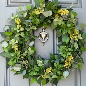 May include: A lush, green wreath composed of various leaves and small yellow flowers. The wreath is hanging on a gray door with a brass door knocker. The door knocker has the text "J. T. Pagliano" and the year "2017" on it.