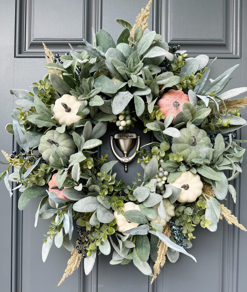 May include: A fall-themed wreath with a variety of greenery, including eucalyptus and lambs ear, accented with small white and pink pumpkins. The wreath is decorated with dried grasses and berries.