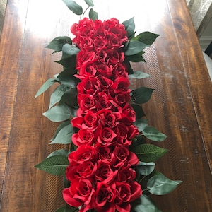 May include: A long, rectangular floral arrangement of red roses and green leaves. The roses are densely packed, creating a vibrant display. The arrangement is placed on a wooden surface, creating a striking contrast.