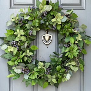 May include: A green and blue wreath with small berries and eucalyptus leaves hangs on a gray door with a silver door knocker. The door knocker has the text "J & T Pagliar" on it.
